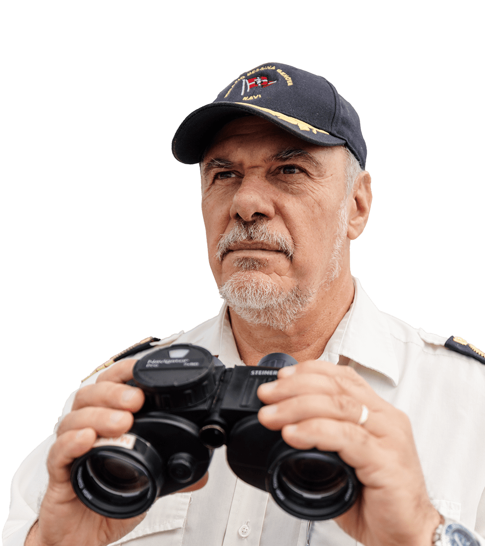 Portrait of the captain of the Jolly Bianco, a middle-aged man with a grey beard wearing the uniform and blue cap with the Messina Line logo. He holds black binoculars and looks into the distance with a serious expression