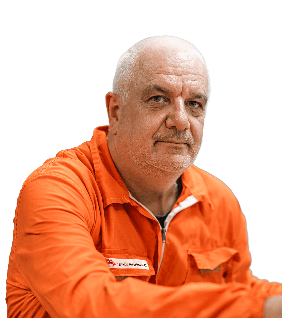 Portrait of a Messina Line crew member in the foreground, he is a mature man with shaved white hair and wears the orange work suit with the logo ’Ignazio Messina & C.’. Look forward with a serene expression