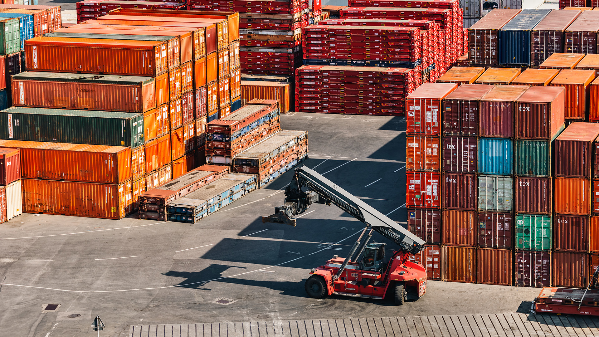 Panoramica dall'alto di un vasto deposito di un terminal container, dominato da alte pile di container marittimi. Un carrello elevatore semovente posizionato nell'area asfaltata tra le pile.