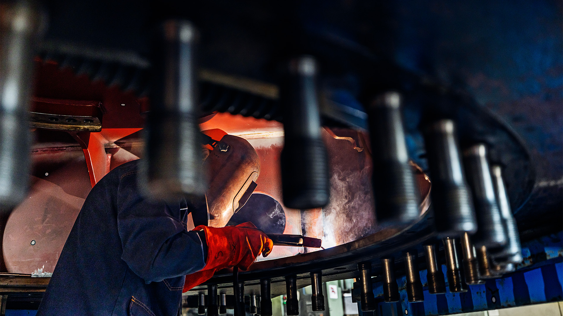 primo piano di un operaio impegnato nella saldatura di un grosso componente metallico.